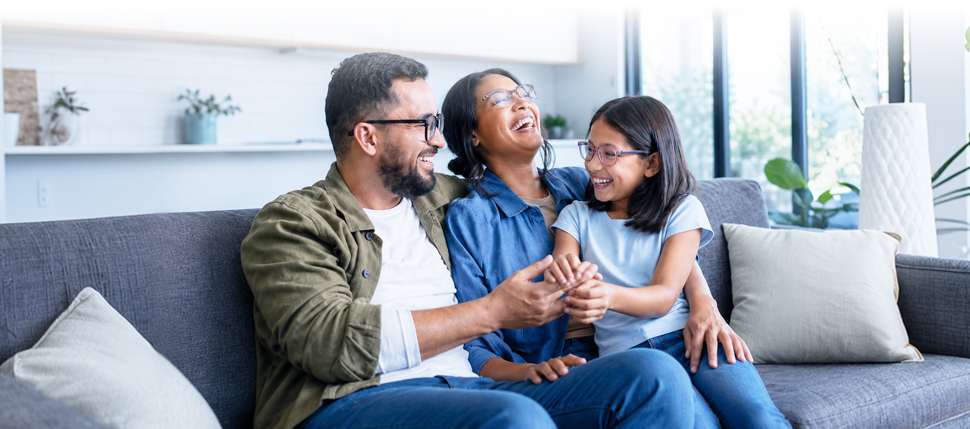 A father, mother, and daughter laugh together on a couch at home.